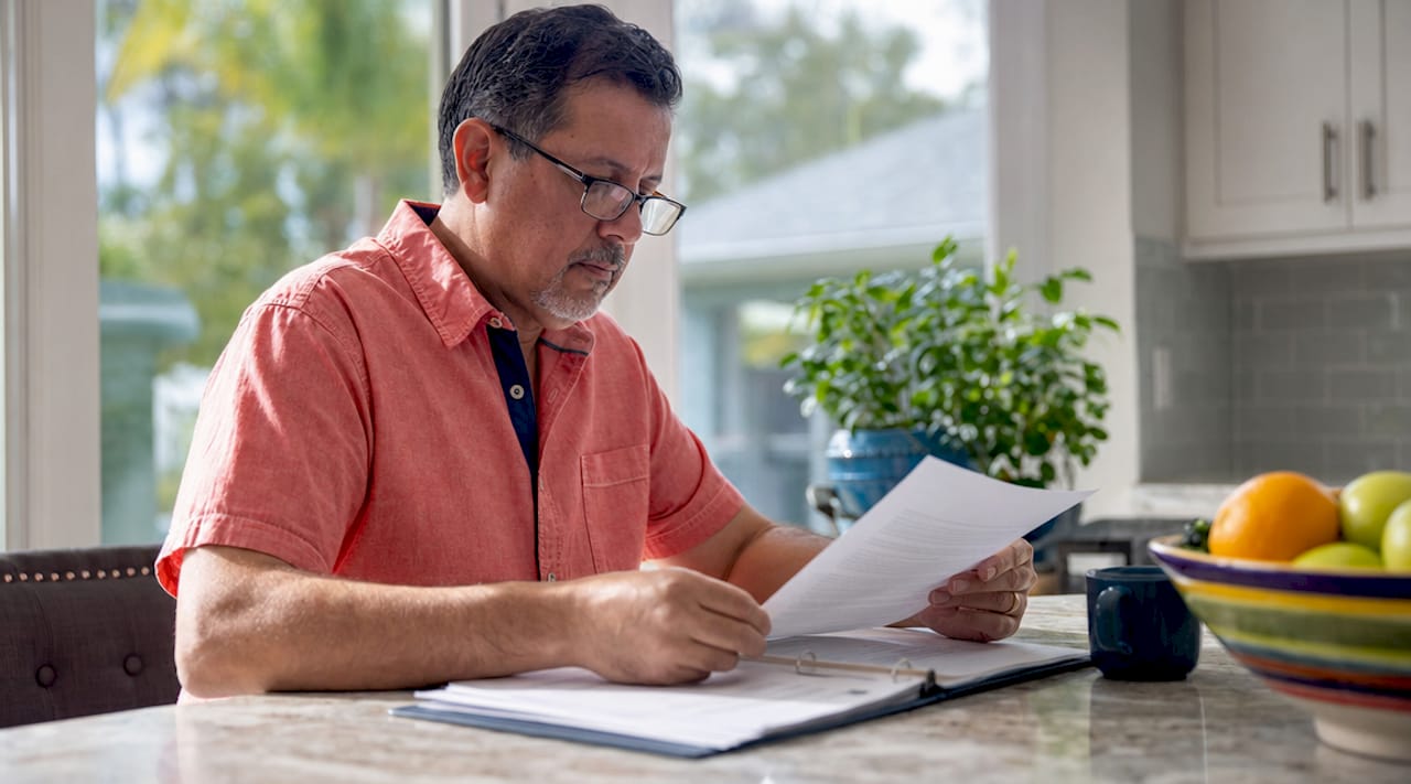 Texas Homeowner in coral shirt and glasses reading an insurance binder document at a bright granite kitchen counter.