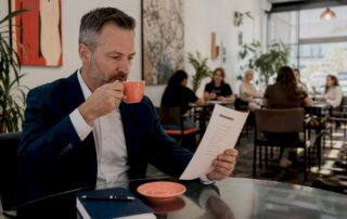 Texas businessman in navy suit sipping coffee while reviewing a printed commercial insurance checklist at a glass café table.