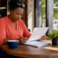 Small business owner in coral henley writing notes on a Texas business personal property inventory spreadsheet at a café table.