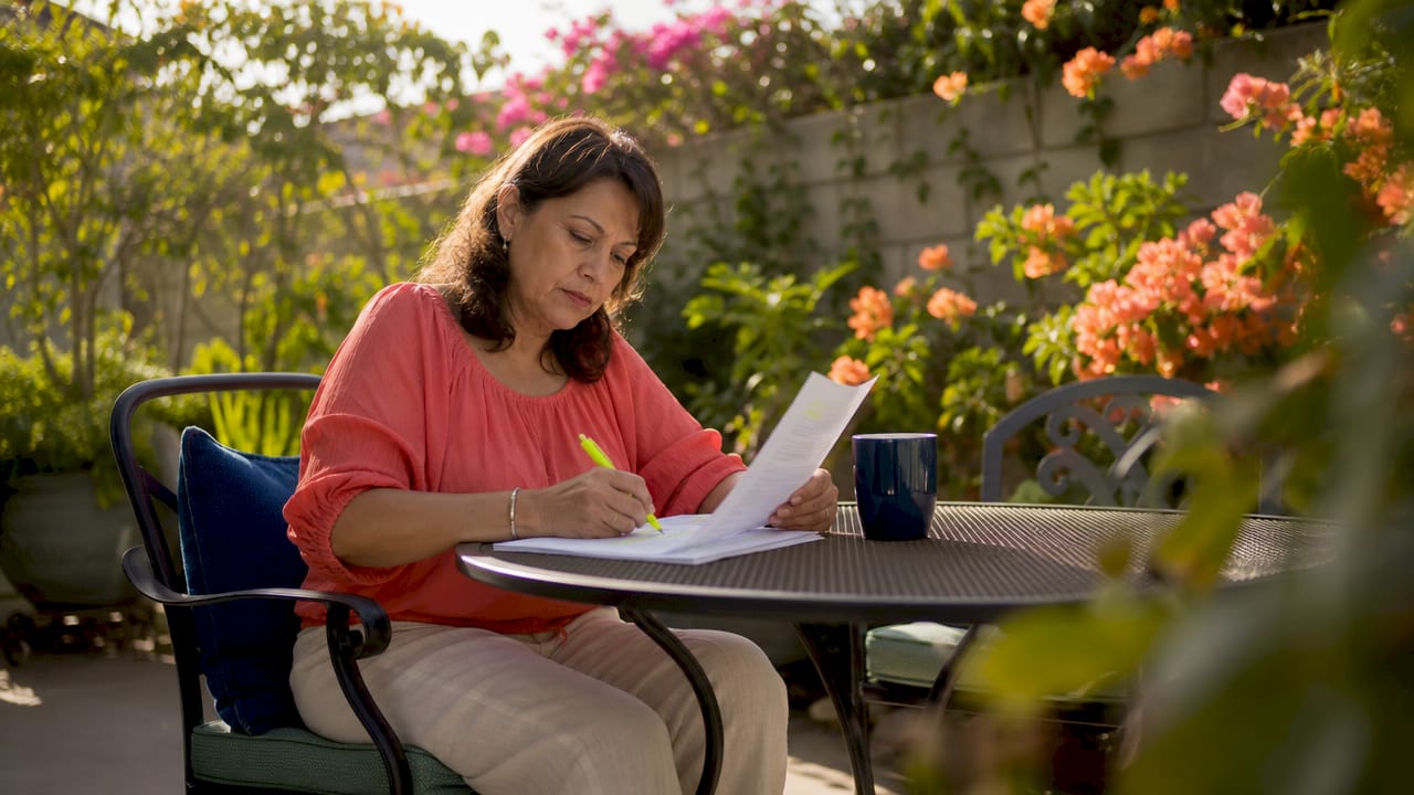 Middle-aged woman in a coral blouse highlighting her Texas insurance policy at a black patio table under coral bougainvillea.
