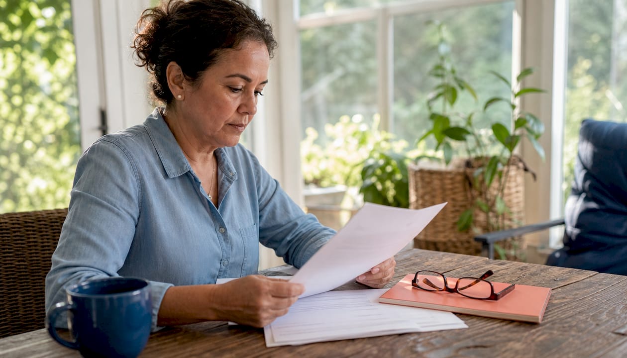 Woman in a chambray shirt reading a Texas insurance declaration page at a wood table with a navy coffee mug and reading glasses.