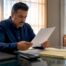 Lubbock businessman in navy shirt reads a commercial insurance document at his glass office desk with laptop and calculator. Looking at Key Coverages for West Texas Businesses, Insurance in 2026.