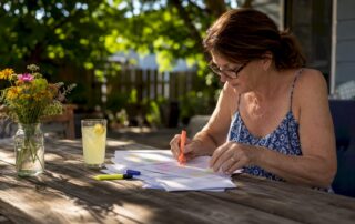 Woman in glasses highlights Texas auto insurance policy pages at a sunny patio table beside lemonade and a jar of wildflowers.