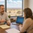 Warm office scene of a male independent insurance agent sitting at a wooden desk reviewing paperwork with a client couple, an open laptop with a blank blue screen and a legal pad with blurred writing on the desk, a coffee mug beside them, and a large window behind showing a flat West Texas landscape under a soft golden late afternoon light.