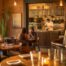 Warm interior of a busy Lubbock Texas restaurant during service with wooden tables, exposed brick, Edison pendant lights, a chef plating food in an open kitchen, and golden afternoon light through a large front window.